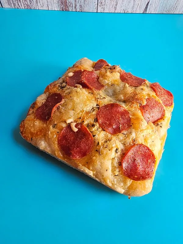 A square sourdough loaf topped with pepperoni, cheese and herbs on a teal board